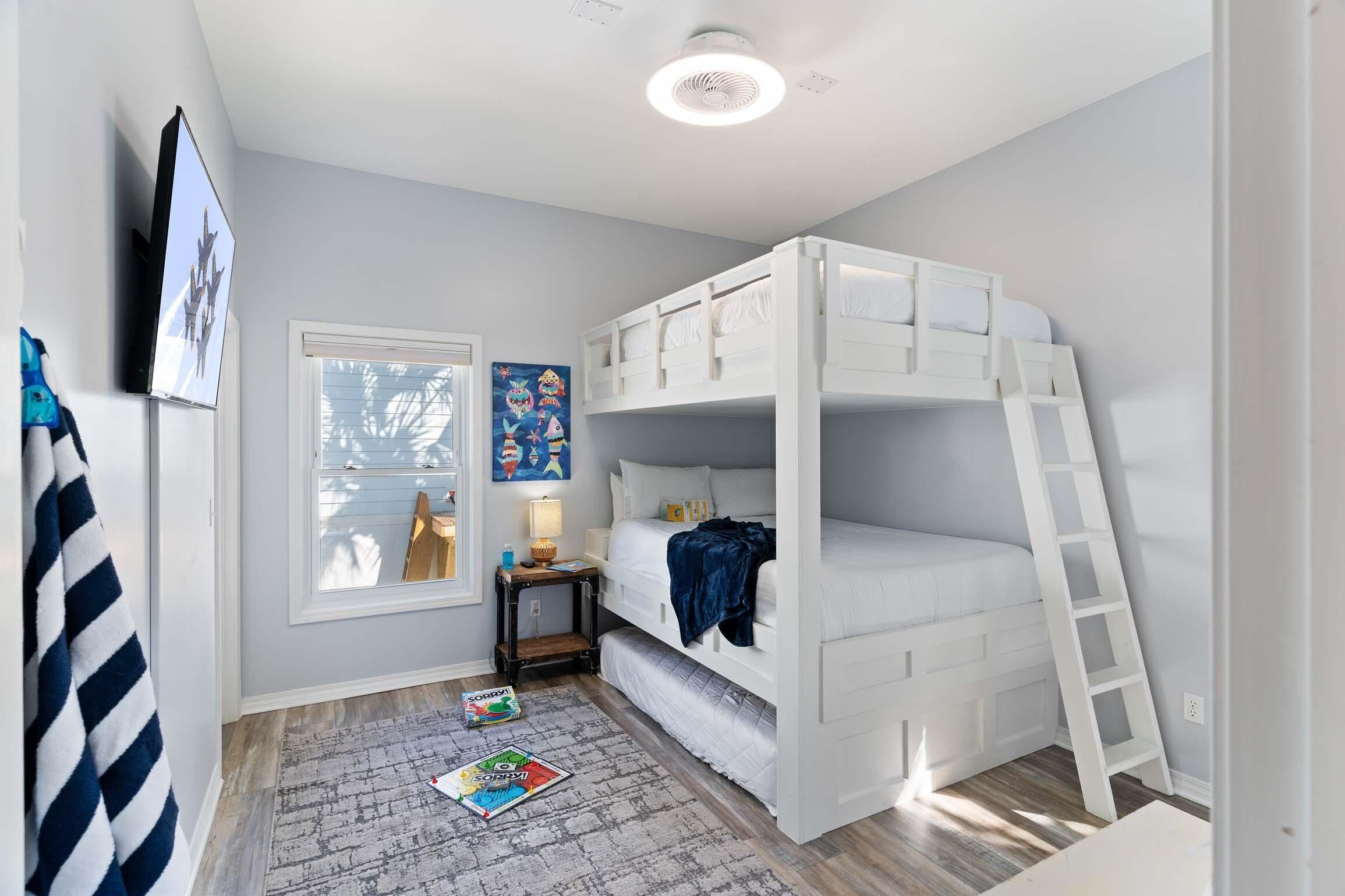 A child's bedroom with bunk beds, a small table, a window with blinds, and a wall-mounted TV.