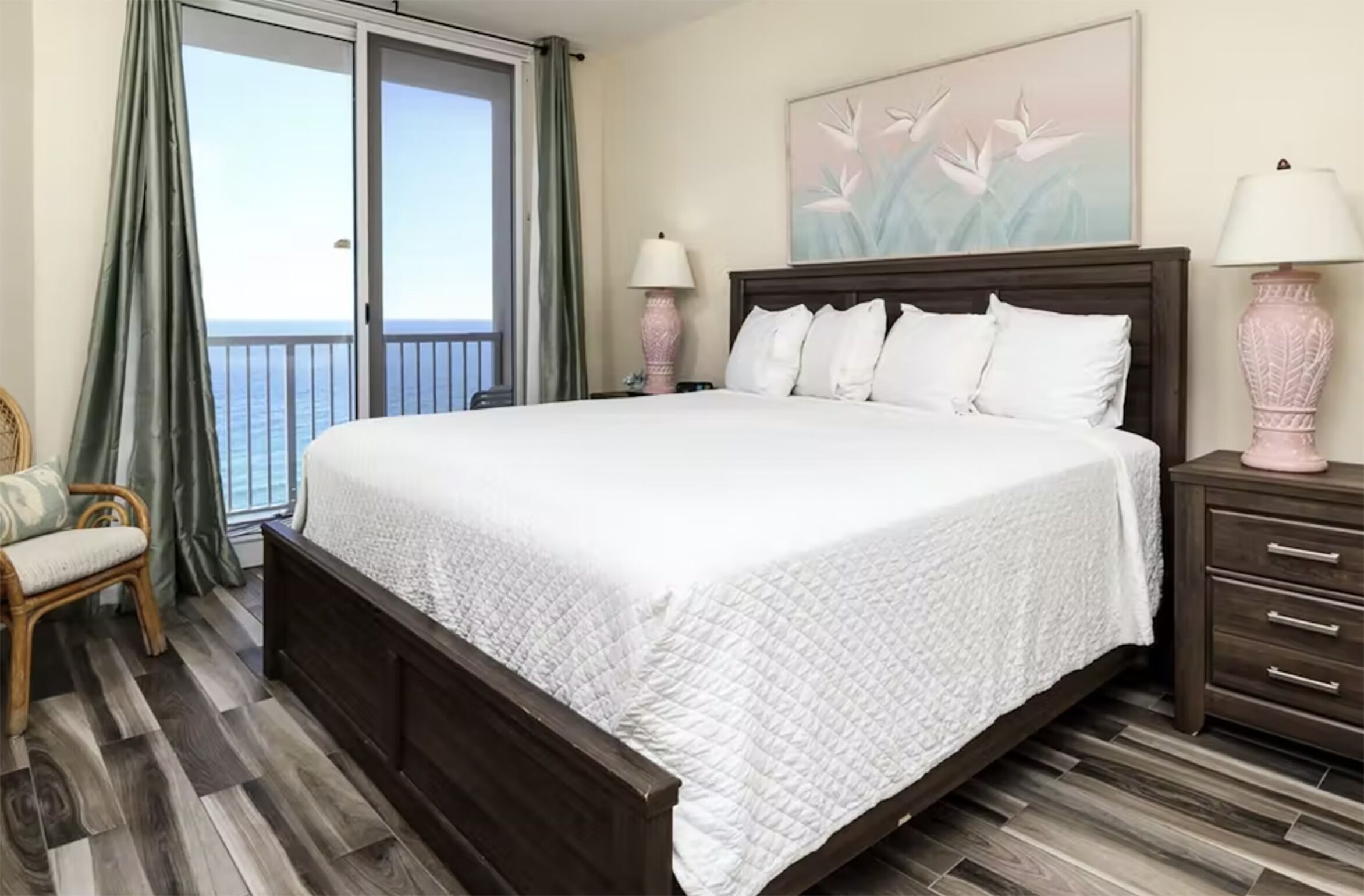 A hotel room with a large bed, a wooden floor, a balcony with curtains, and two bedside lamps.
