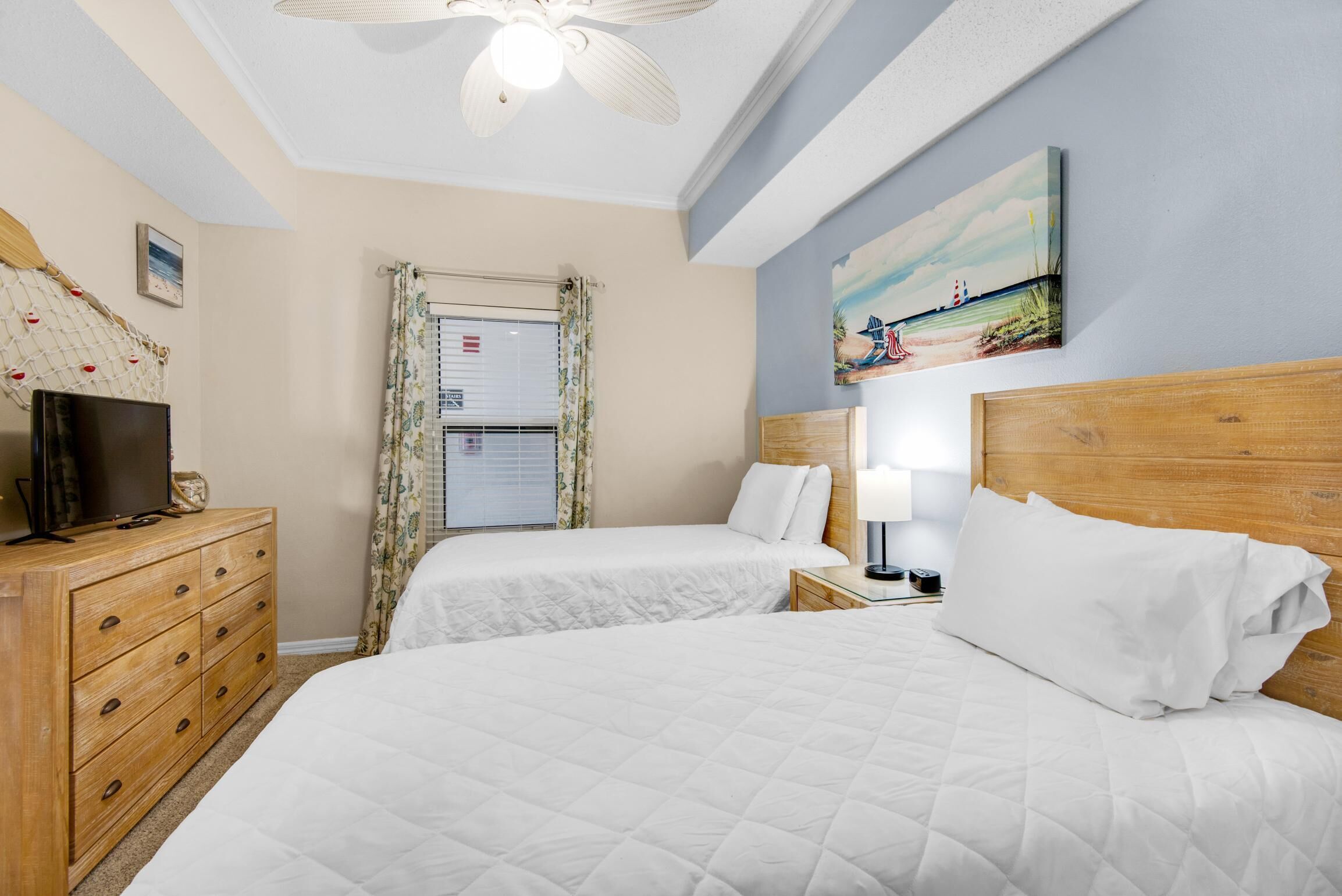 A hotel room with two beds, a wooden dresser, a television, a ceiling fan, a window with curtains, and a beach-themed painting on the wall.