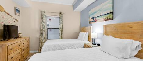 A hotel room with two beds, a wooden dresser, a television, a ceiling fan, a window with curtains, and a beach-themed painting on the wall.
