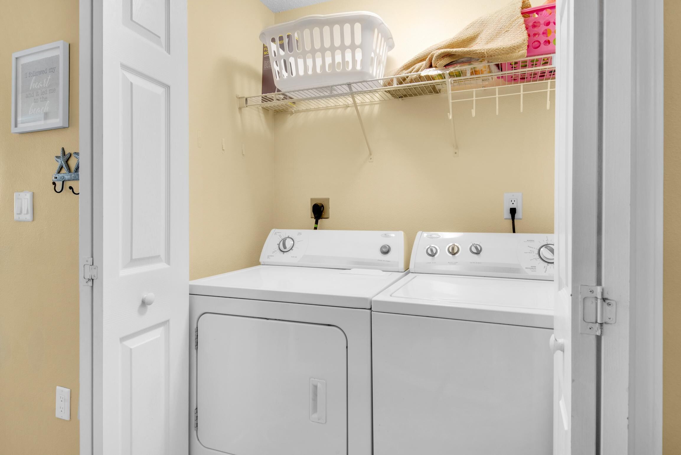 Une buanderie équipée de deux machines à laver et de deux sèche-linge à chargement frontal, tous deux blancs, d’un panier à linge blanc et d’une étagère où sont pliées des serviettes.