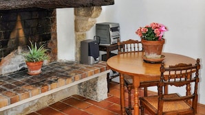 Un interior rústico con chimenea de piedra, una mesa de madera y sillas. Hay una maceta sobre el alféizar de la chimenea y otra sobre la mesa.