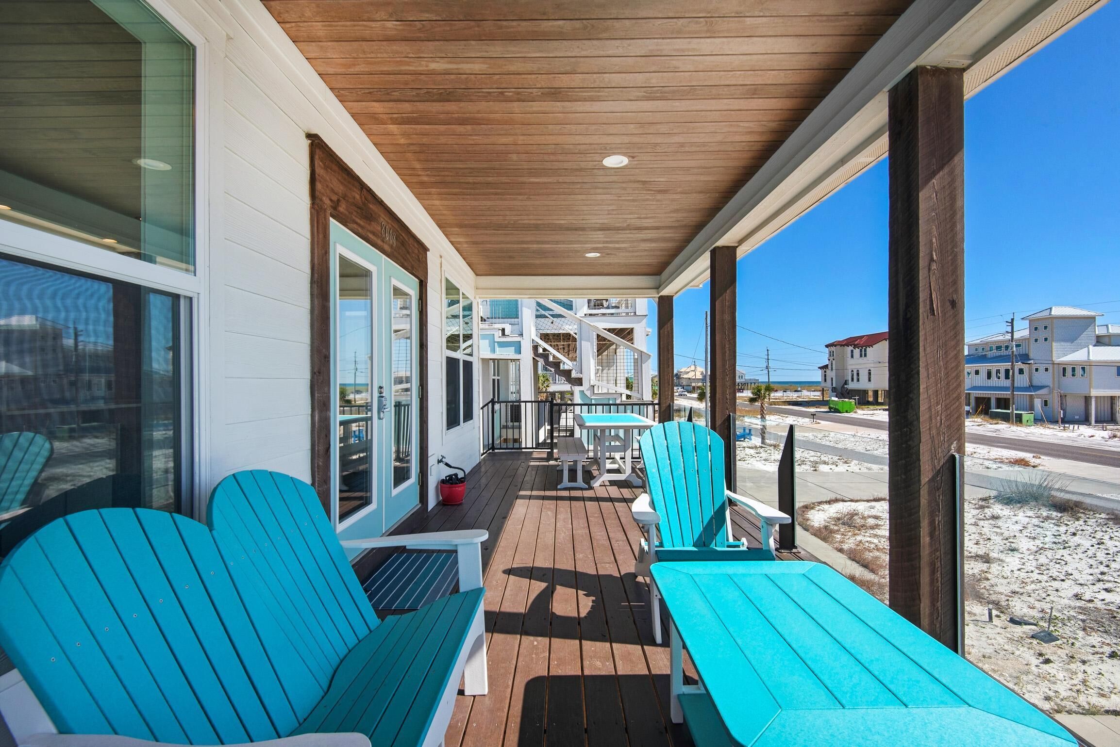 Un espace extérieur abrité, doté d’un plancher en bois, de chaises et de tables bleues, et offrant une vue sur une plage et des bâtiments.