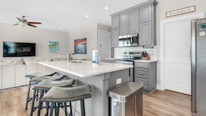 A modern kitchen with grey cabinets, a large island with bar stools, a ceiling fan, and a sign that reads 'BEACH HOUSE'. - Para-dise Upper (Navarre)