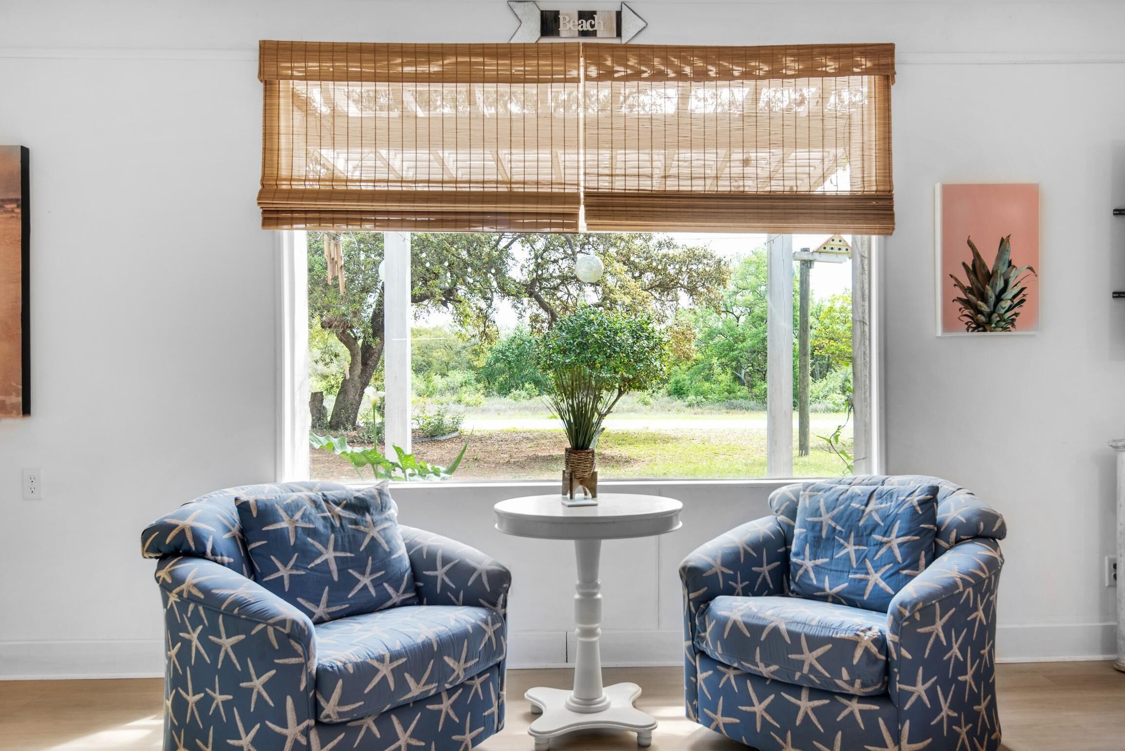 Ein Zimmer mit zwei blauen Sesseln, einem kleinen weißen Tisch, einem Fenster mit Blick auf Grünflächen und einem an der Wand befestigten Schild mit der Aufschrift 'Beach'.