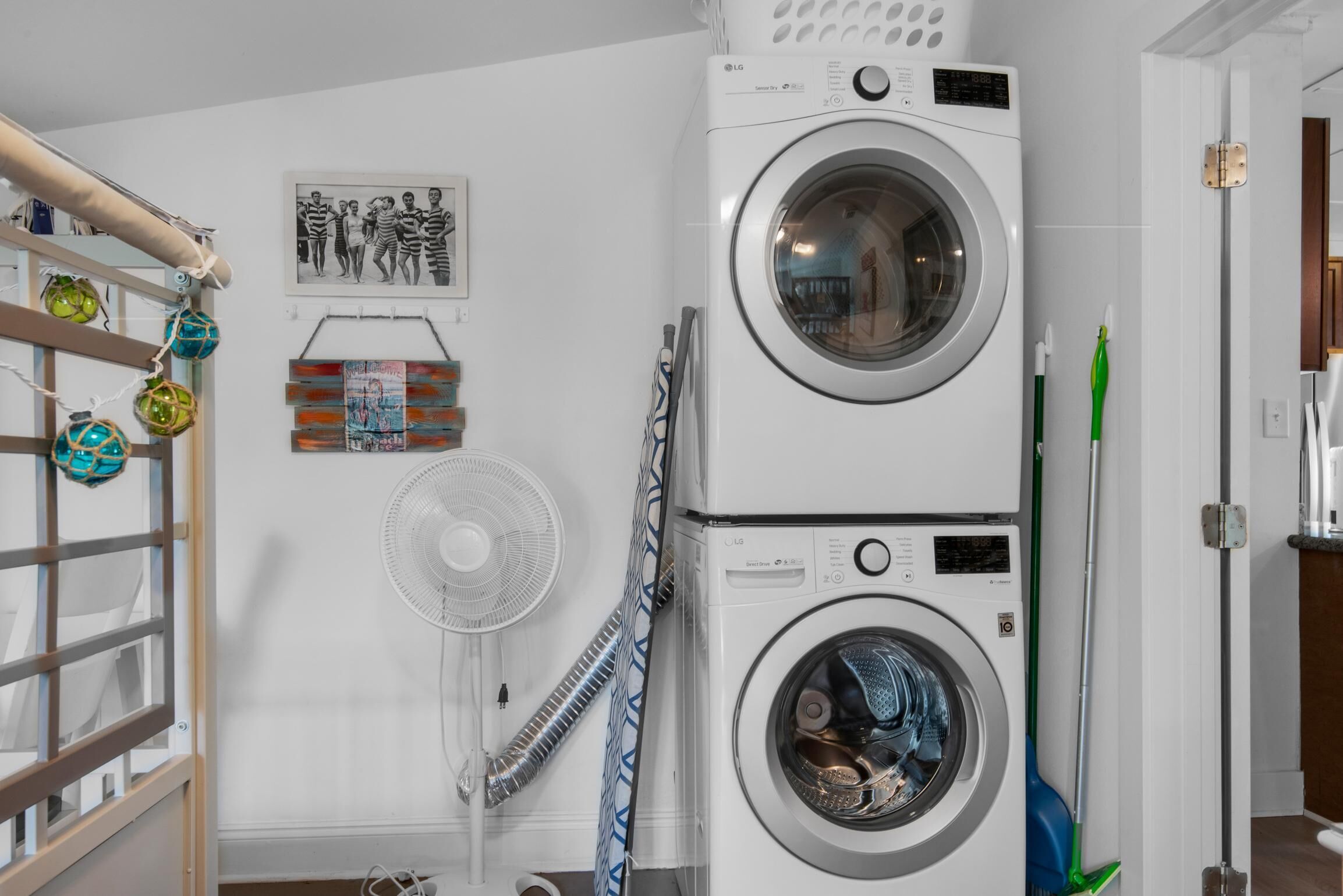 Un espace dédié à la buanderie, équipé de machines à laver et de sèche-linge superposés, d’un ventilateur et d’un balai.