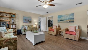 A living room with a ceiling fan, a white coffee table, two wicker chairs, a bookshelf, and a painting on the wall. - Bungalow By the Sea-starfish (Navarre)