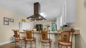 A kitchen with a central island, three wicker chairs, a stainless steel range hood, and a refrigerator. - Bungalow By the Sea-starfish (Navarre)