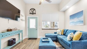 A living room with a blue sectional sofa, a teal console table, a flat-screen TV, and a door with a sign above it. - Para-dise LZ (Navarre)