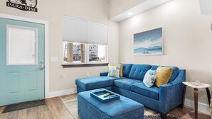 A living room with a blue sectional sofa, a small side table, and a window with blinds. - Para-dise LZ (Navarre)