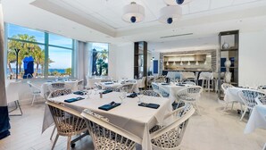 A spacious dining area with multiple tables and chairs, large windows offering a view of palm trees and the sea, and modern pendant lighting. - Portofino Island Resort 5-1708 (Pensacola Beach)