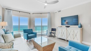 A spacious living room with a large glass coffee table, blue armchairs, a white sofa, and a television on the wall. - Emerald Isle Unit 703 (Pensacola Beach)