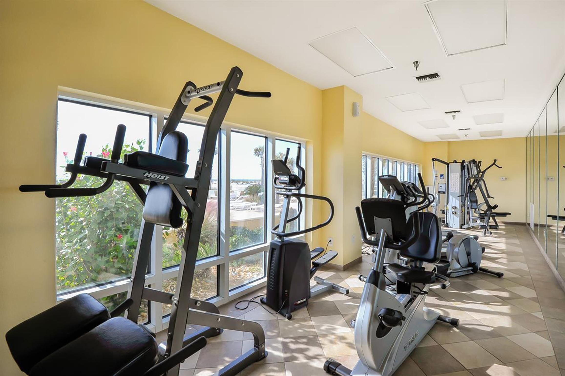 Une salle de sport équipée de divers appareils d’exercice, dotée de grandes fenêtres et offrant une vue sur la verdure environnante.