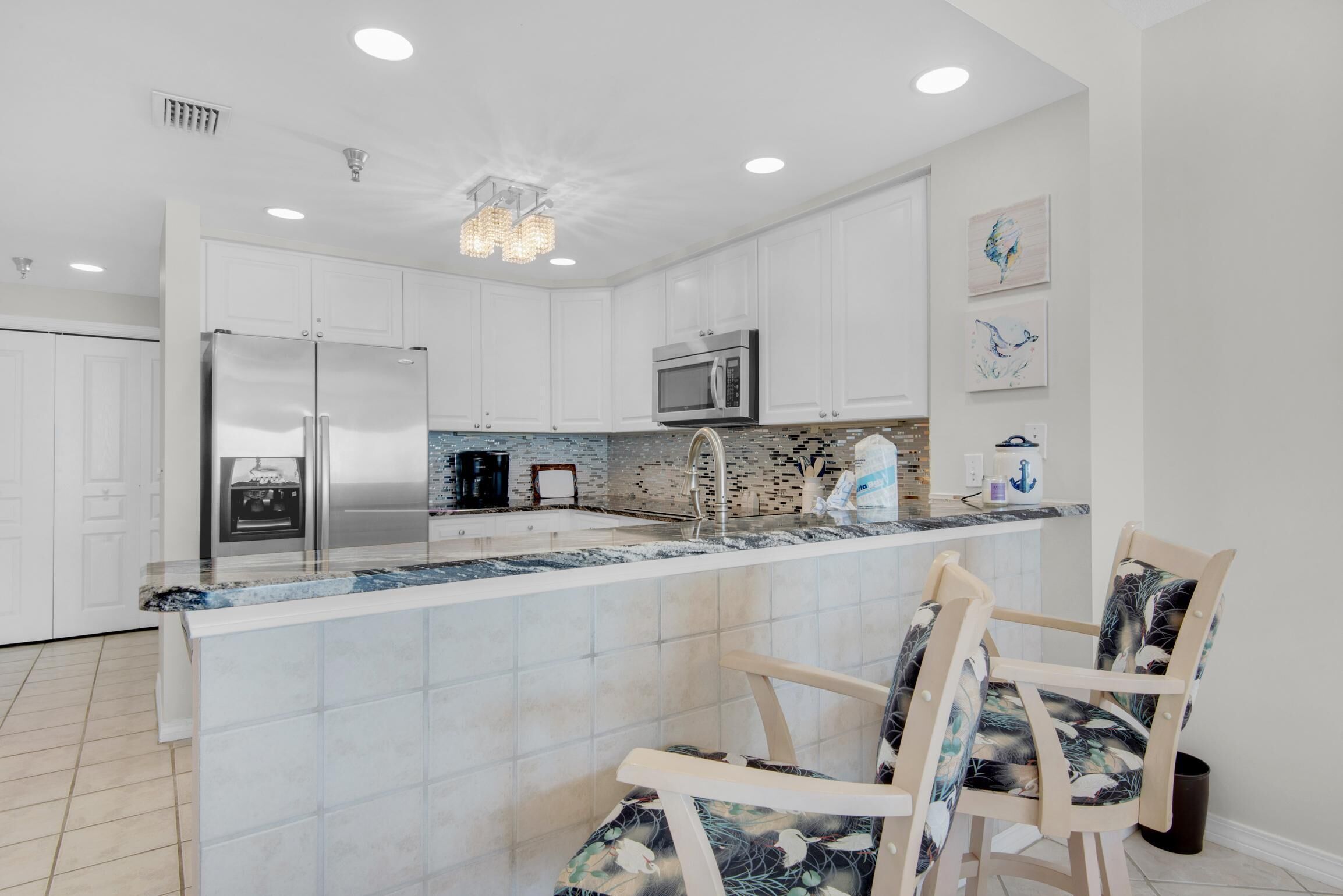 A modern kitchen with white cabinetry, stainless steel appliances, a marble countertop, and a small island with two chairs.