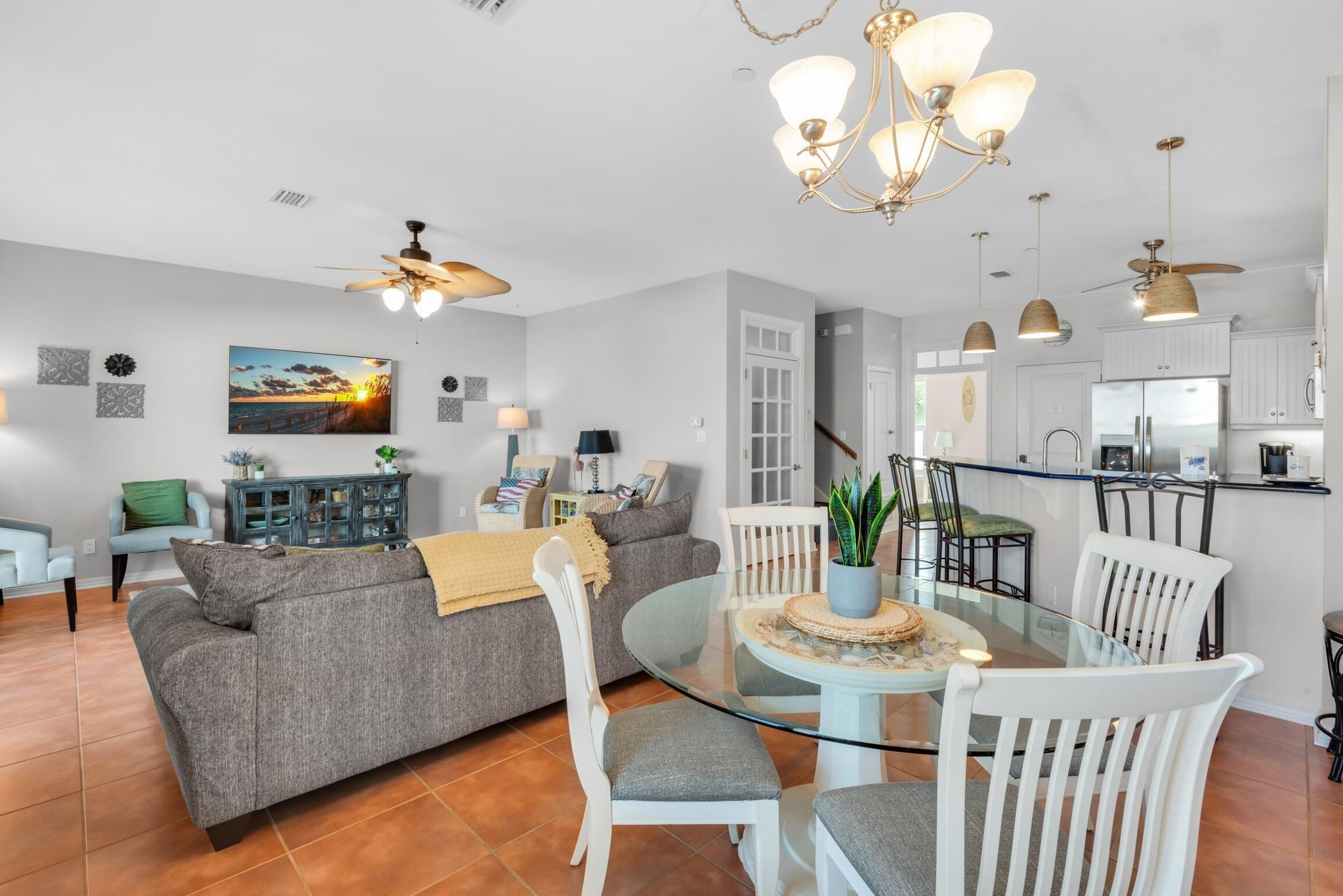 Un salon moderne avec une table à manger en verre, des chaises blanches, un canapé gris et un ventilateur de plafond.