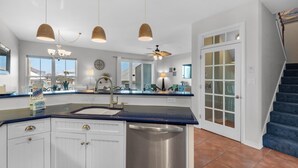 A modern kitchen with white cabinets, a stainless steel dishwasher, and a blue countertop. There are three pendant lights, a ceiling fan, and a large window providing natural light. - Bonita Bay (Pensacola Beach)