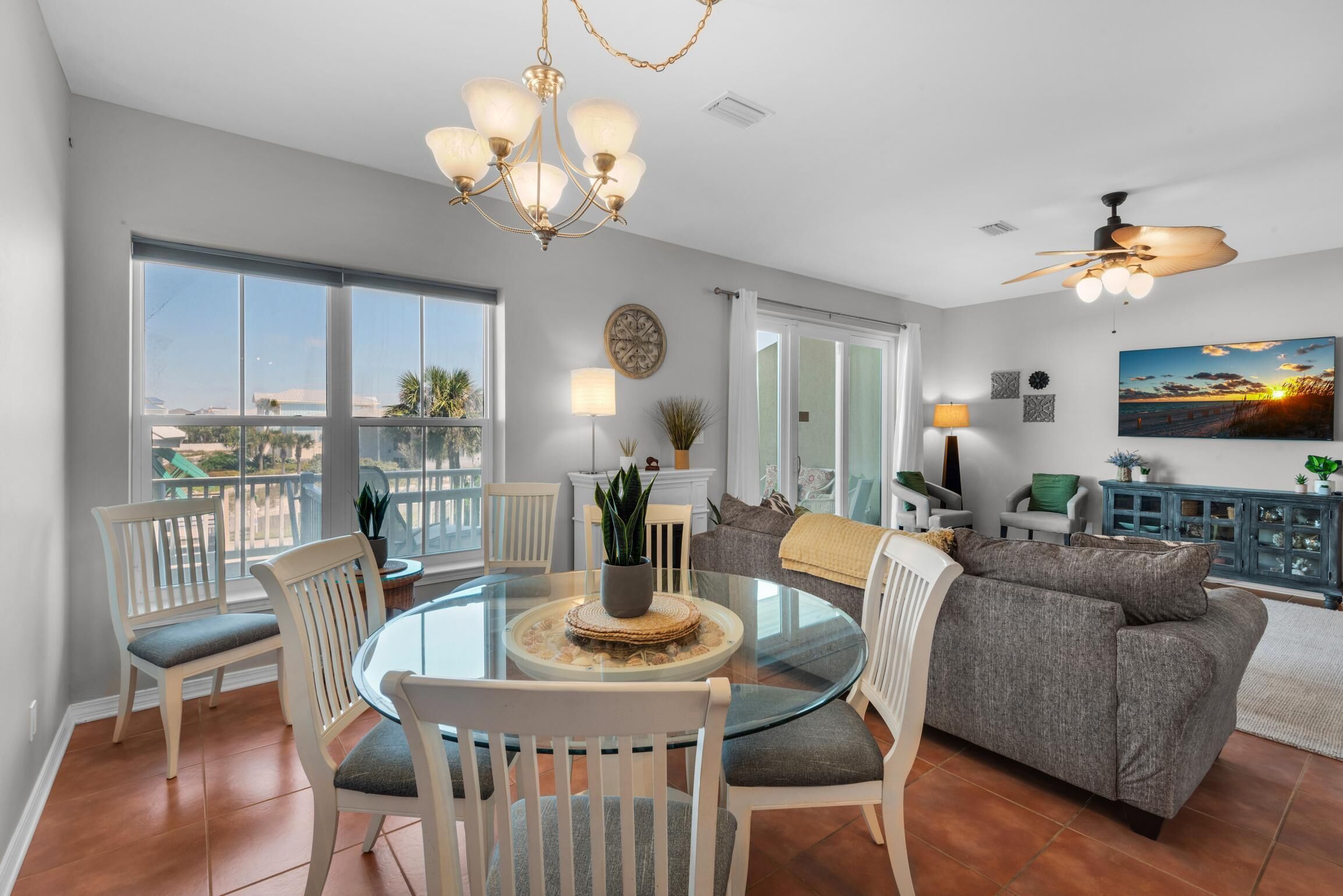 A spacious living room with a glass dining table surrounded by white chairs, a grey sofa, and a ceiling fan.