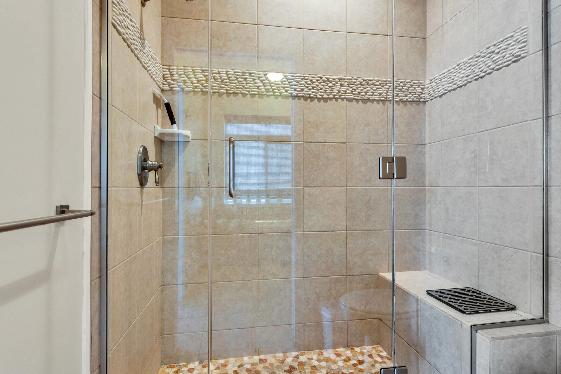 Une douche fermée par des parois en verre, avec des carreaux de couleur beige, une barre d’appui en métal et une petite étagère dotée d’un drain.