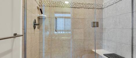 A glass-enclosed shower with beige tiles, a metal handrail, and a small shelf with a drain.