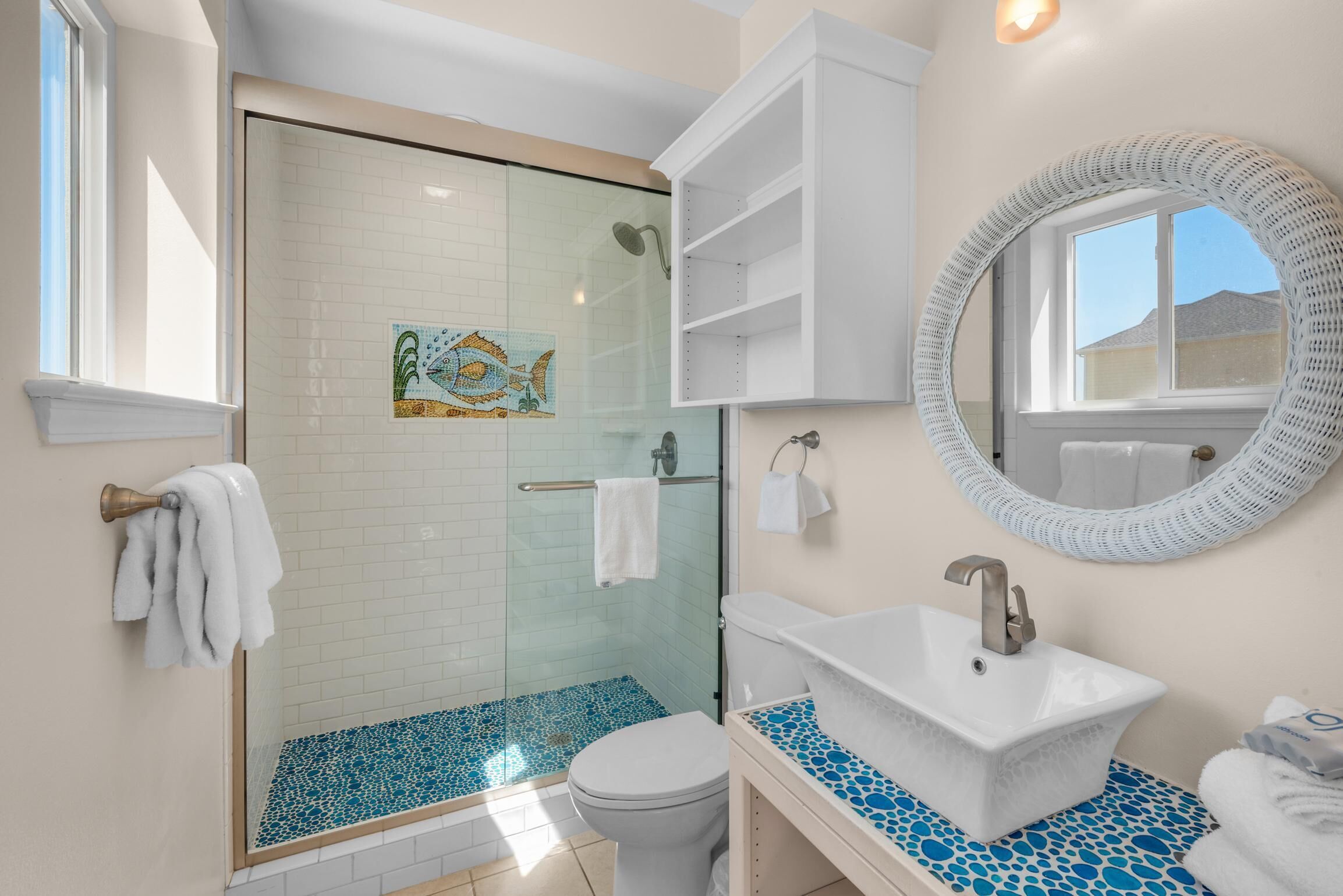 Un baño con ducha vidriada, un lavamanos blanco con encimera de mosaico azul y un espejo redondo sobre el lavamanos.