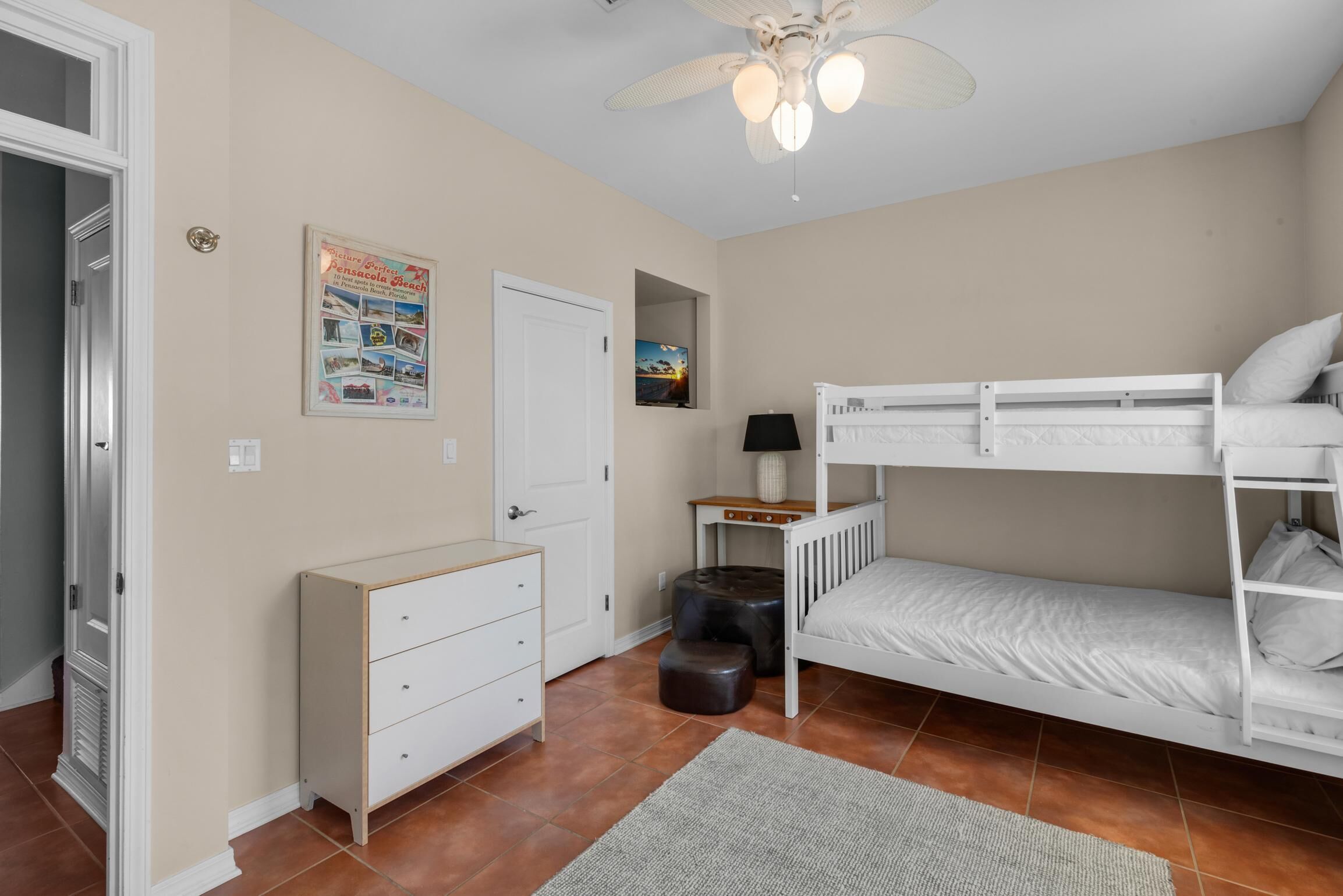 Een kamer met een stapelbed, een bureau met een lamp, een dressoir, een plafondventilator en een lijst aan de muur.