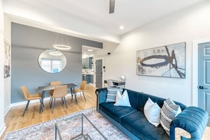 Ein modernes Wohnzimmer mit einem blauen Sofa, einem Essbereich mit einem runden Tisch und einer Küche im Hintergrund.