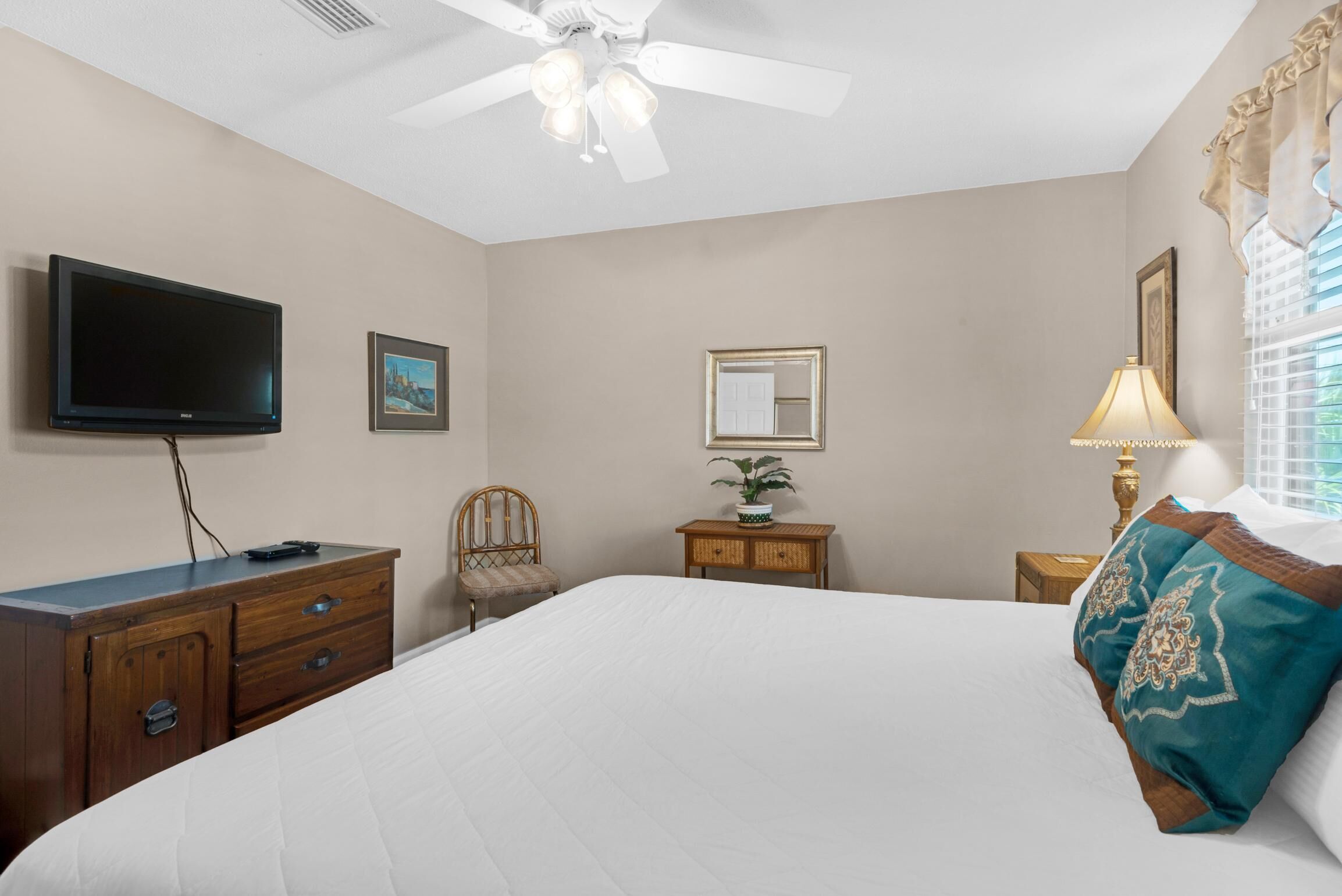 A bedroom with a bed, a television, a ceiling fan, a dresser, a chair, a nightstand, a lamp, and a window with curtains.