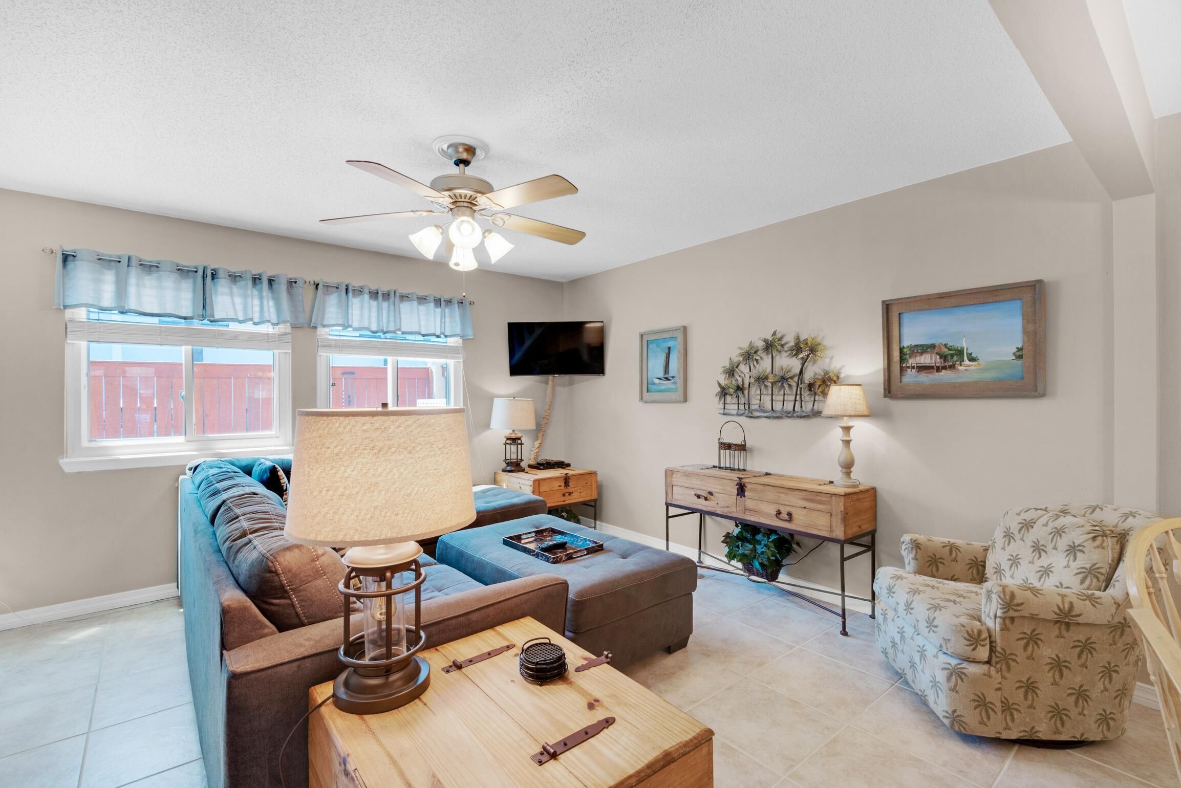 A living room with a sofa, ottoman, coffee table, and a chair. There is a ceiling fan, a TV mounted on the wall, and a window with curtains.