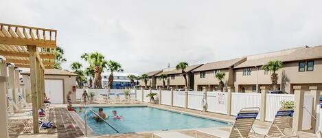 A pool area with lounge chairs, a covered seating area, and a building with multiple windows and a flat roof.