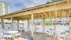 An outdoor patio area with white tables and chairs, a wooden pergola, and a view of residential buildings in the background. - Sugar Beach F-27 (Navarre)