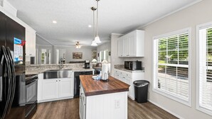 A modern kitchen with white cabinets, a wooden island, a black trash can, and a window with blinds.