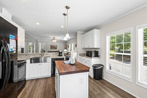 A modern kitchen with white cabinets, a wooden island, a black trash can, and a window with blinds. - Beachin' Fun (Navarre)