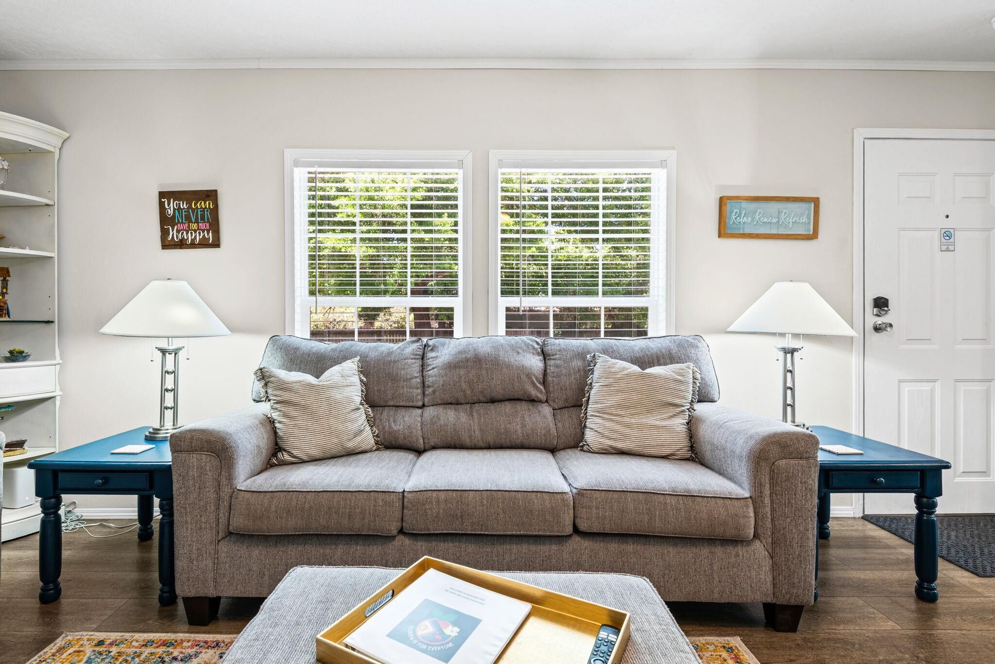 A living room with a grey sofa, two side tables with lamps, a coffee table with a tray, and two windows with blinds.