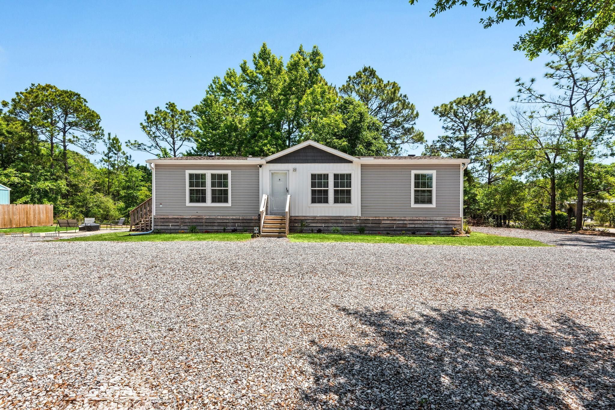 A single-story mobile home with a front porch, a gravel driveway, and a wooden deck.