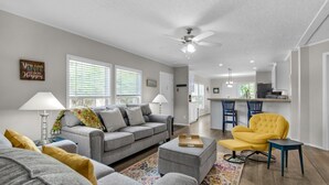 A living room with a gray sofa, a yellow armchair, and a coffee table. There is a kitchen in the background with bar stools.