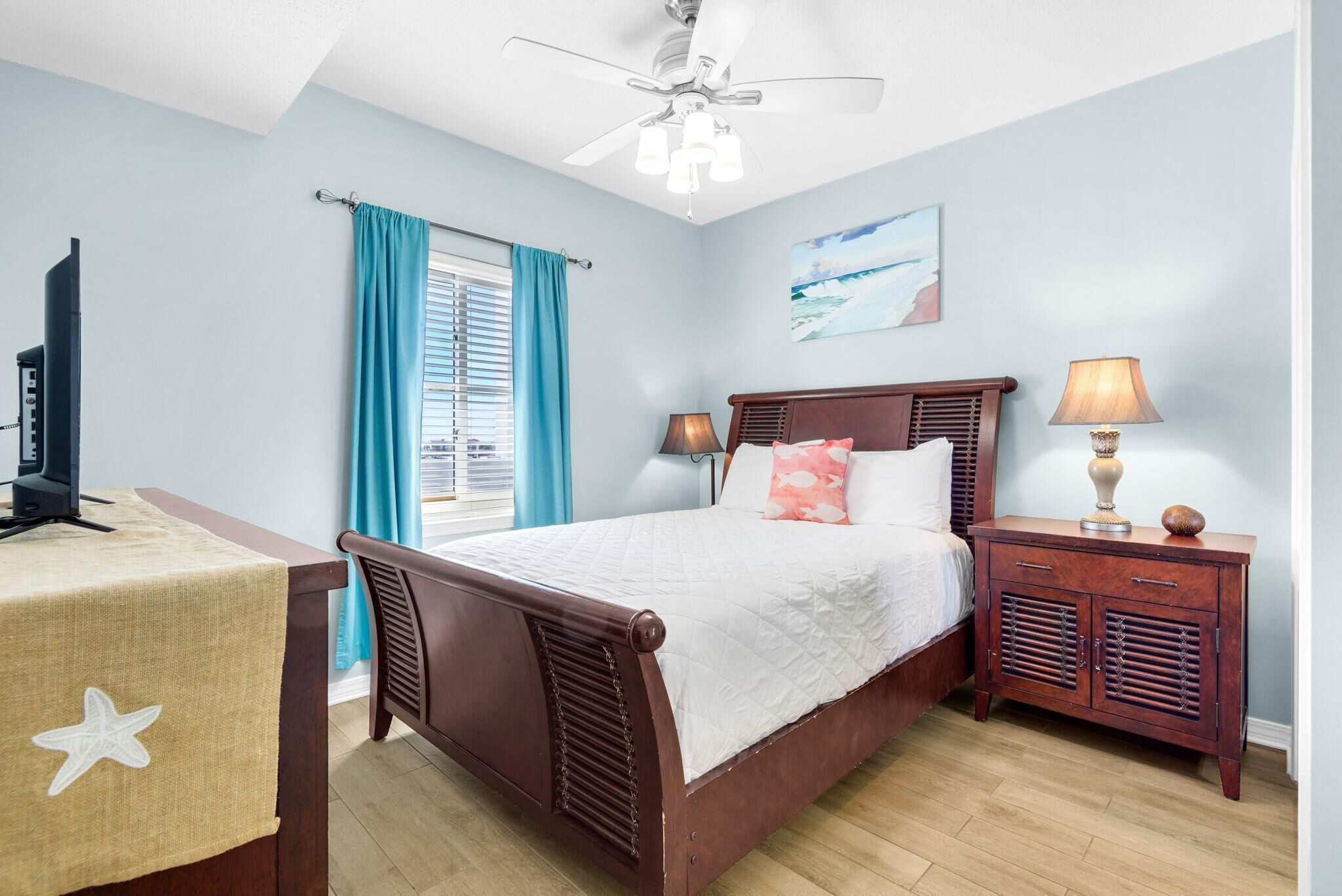 A bedroom with a bed, a desk, a nightstand, a ceiling fan, and a window with curtains.