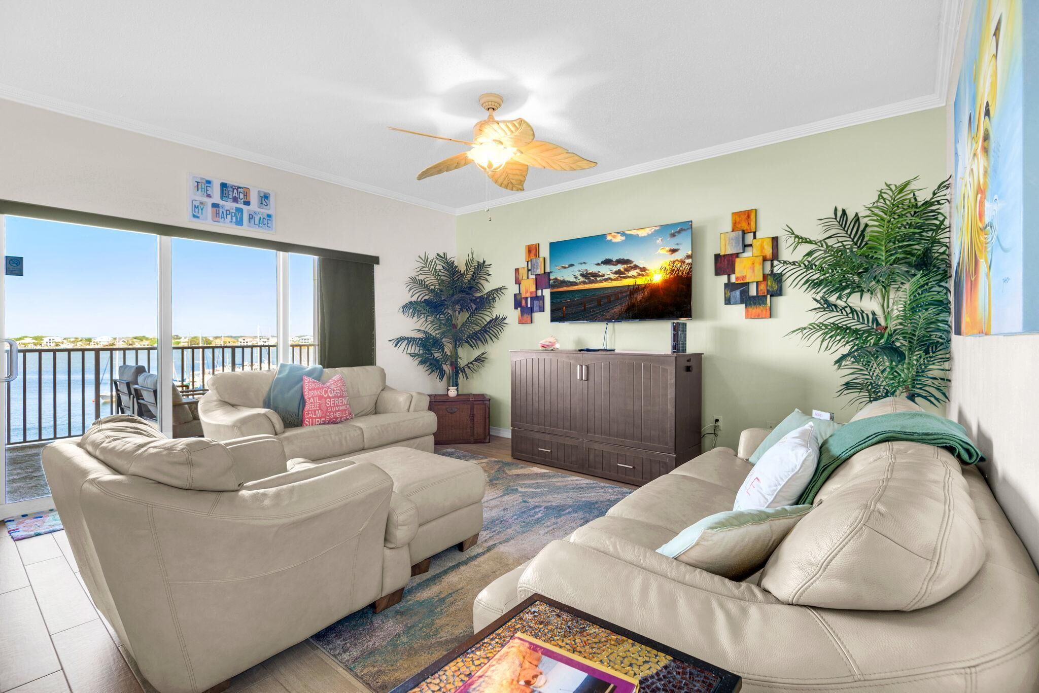 A living room with a sofa set, a TV, a ceiling fan, a plant, and a view of the beach.