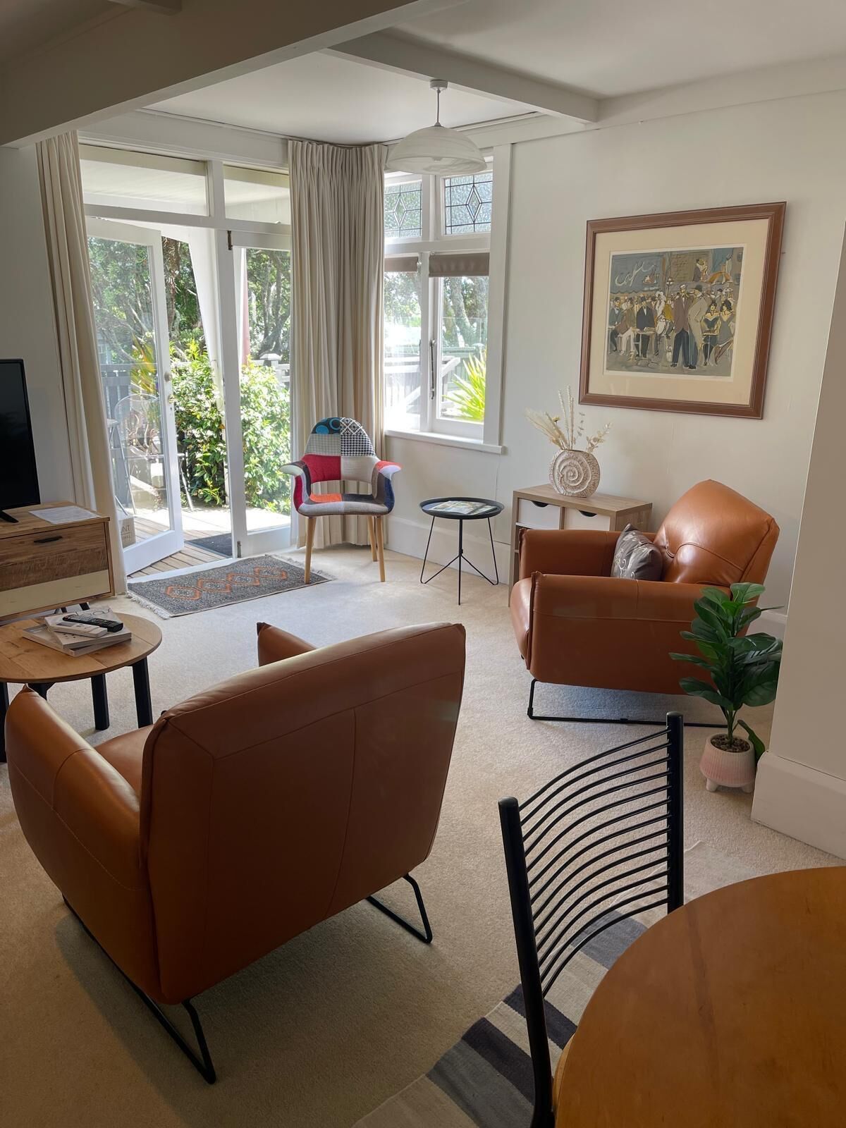 A modern living room with a sofa, armchairs, a coffee table, and a TV. There is a large window with curtains, a painting on the wall, and a small table with a vase.