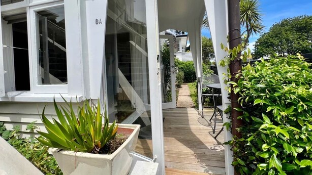 Uma casa branca com deck de madeira, porta de vidro e um vaso com planta em primeiro plano.