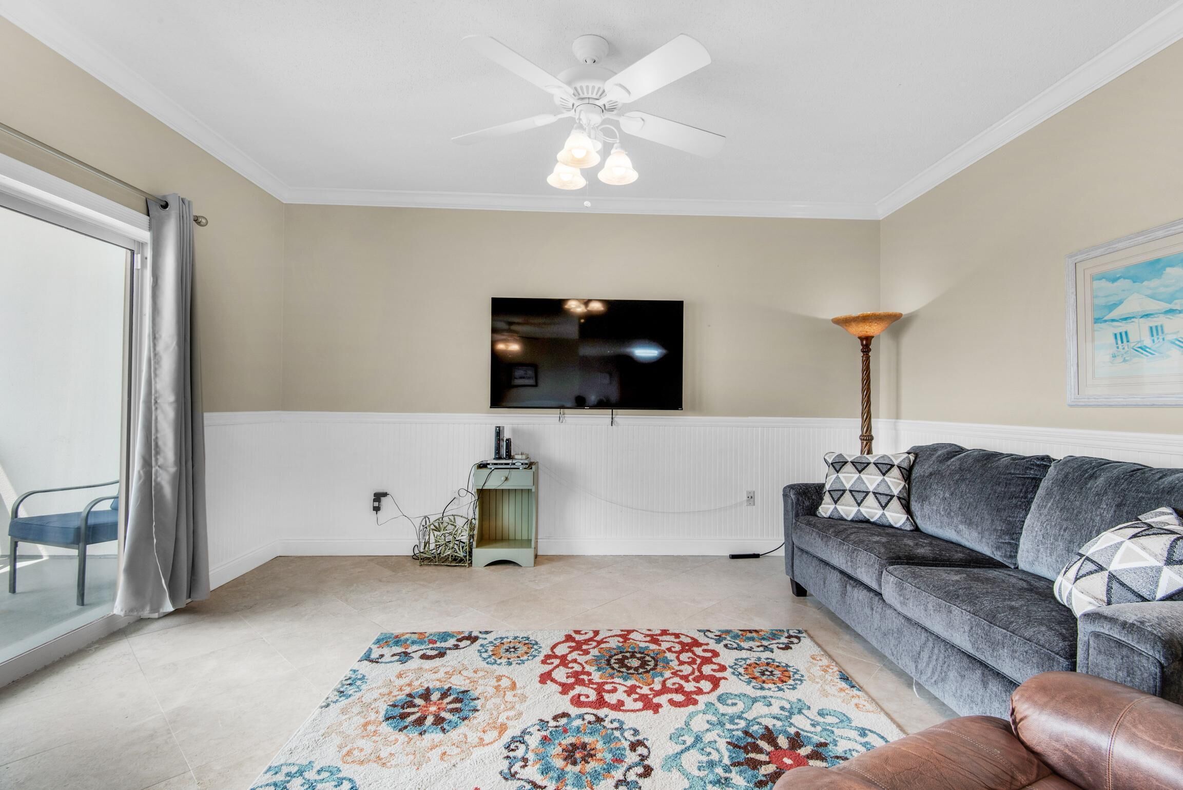 A living room with a ceiling fan, a flat-screen TV mounted on the wall, a patterned rug, a gray sofa, and a brown leather chair.