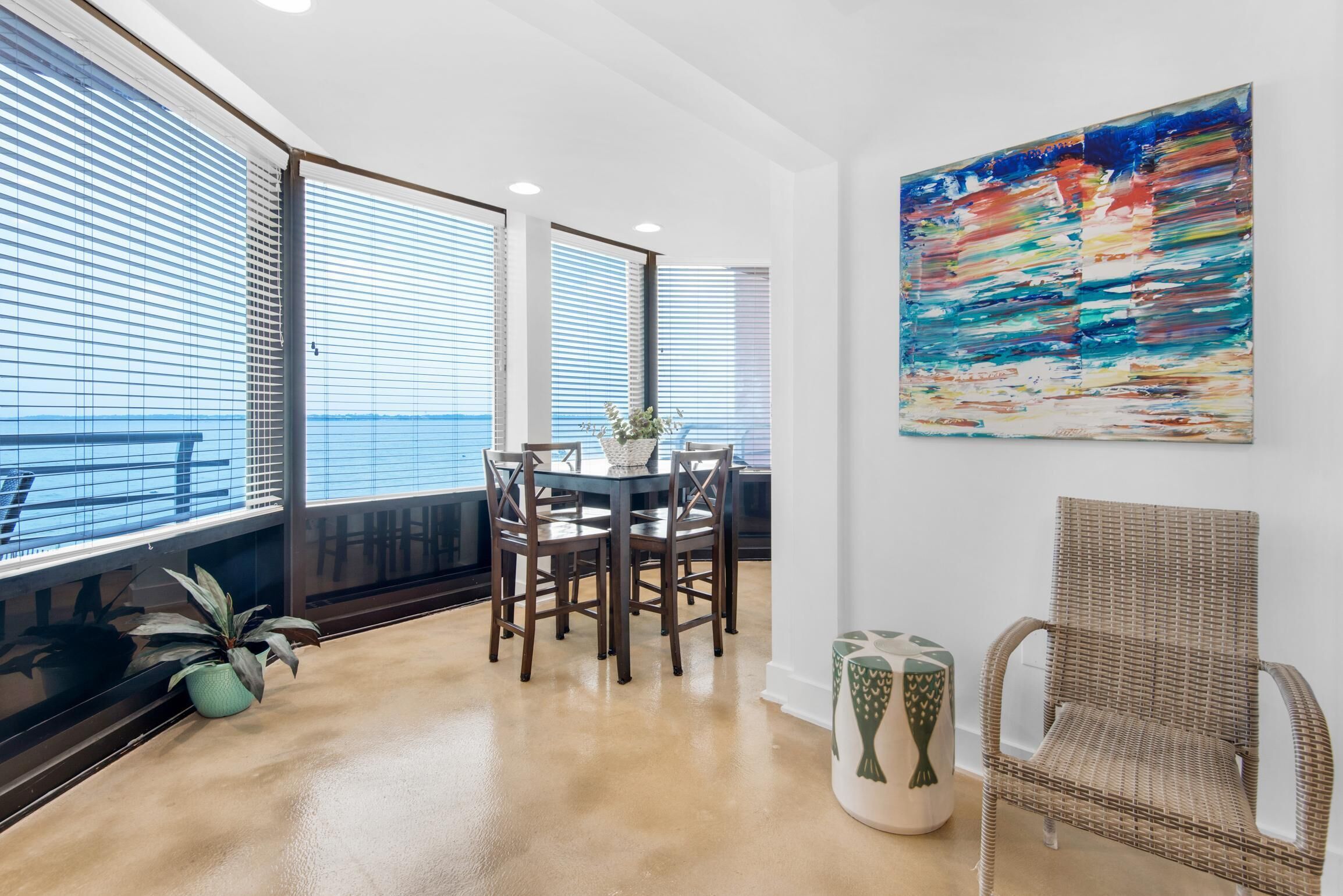 A modern dining area with a round table and chairs, large windows with blinds, a wicker chair, and a colorful abstract painting on the wall.