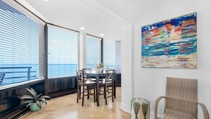 A modern dining area with a round table and chairs, large windows with blinds, a wicker chair, and a colorful abstract painting on the wall. - Palm Beach Club 2-112 The Sugar Spot (Pensacola Beach)