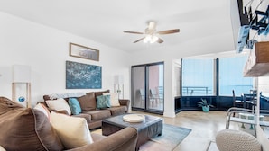 A spacious living room with a brown sofa, a coffee table, and a ceiling fan. There is a large window with a view of the outdoors. - Palm Beach Club 2-112 The Sugar Spot (Pensacola Beach)