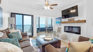 A spacious living room with a large window, a ceiling fan, a flat-screen TV, and a fireplace. - Palm Beach Club 2-112 The Sugar Spot (Pensacola Beach)
