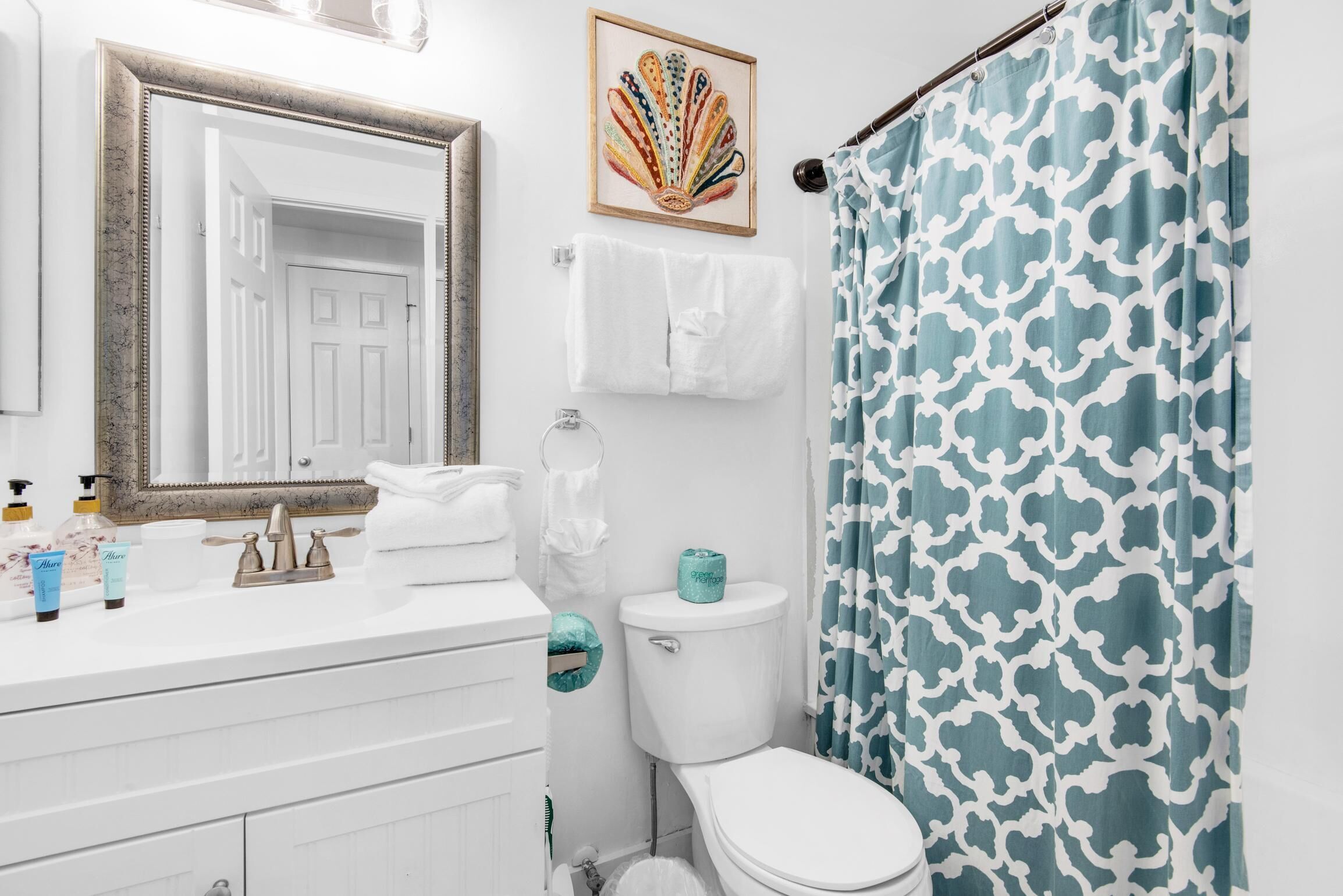 Un baño con tocador blanco, un espejo grande, un inodoro y una cortina de ducha con estampado azul y blanco.
