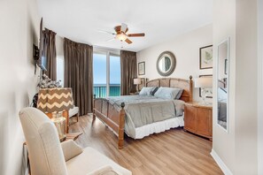 Une chambre avec un lit, un ventilateur de plafond, une table de chevet, une chaise, une lampe, un miroir et une vue sur l’océan.