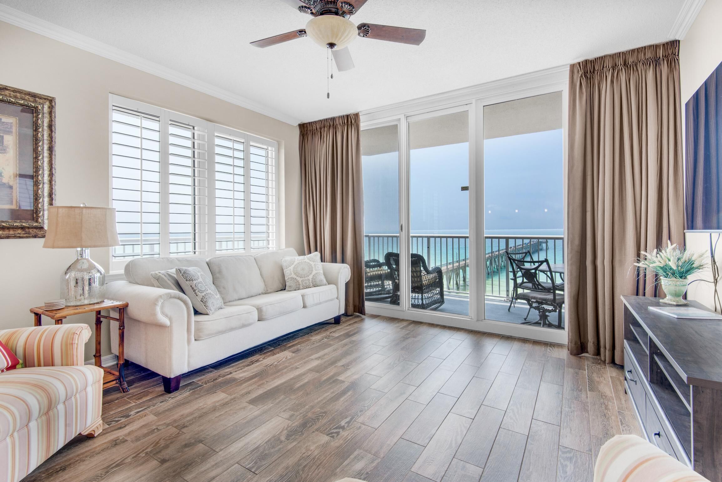 A spacious living room with a white sofa, a wooden floor, a ceiling fan, and a view of the ocean through large sliding doors.