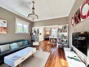 Uma sala de estar ampla com um sofá grande, uma mesa de centro e uma TV. Estantes e um espaço para refeições são visíveis ao fundo.