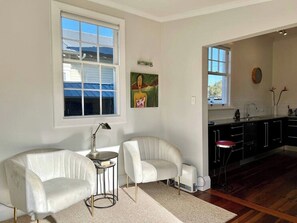 Uma sala moderna com um sofá branco, uma pequena mesa lateral e um quadro na parede. Há uma área de cozinha com armários escuros e uma janela.