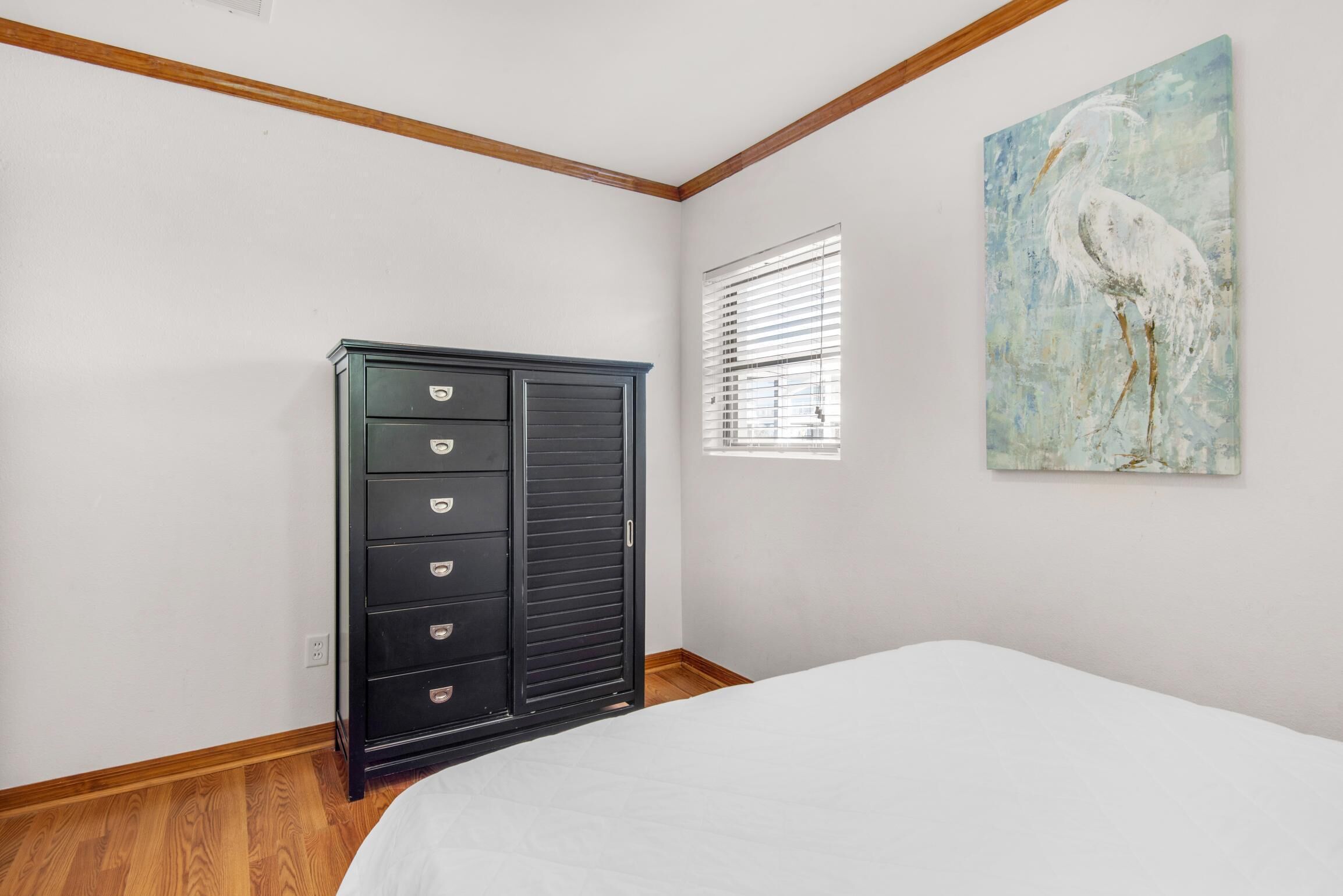 A bedroom with a bed, a dresser, a window with blinds, and a wall painting of a bird.